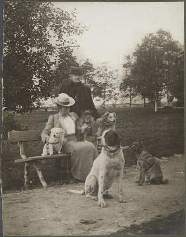 Kaksi naista puistossa. Toinen istuu penkillä, toinen seisoo hänen takanaan. Heidän kanssaan on viisi koiraa.