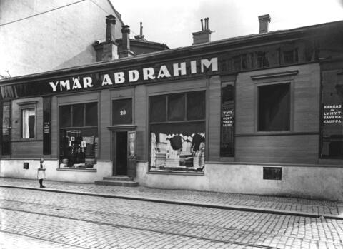 I förgrunden en kullerstensgata, en flicka med flätor står framför en låg träbyggnad som inhyser en butik.