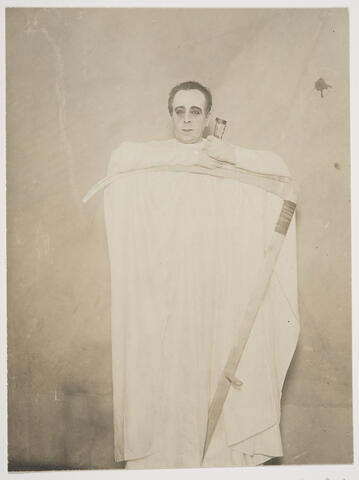 Valkoiseen huputtomaan kaapuun pukeutunut voimakkaasti maskeerattu mies seisoo nojaamassa viikatteen terään.