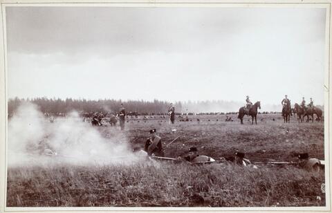Men and horses during military exercises in the field. One man is lying on the ground and firing a rifle.
