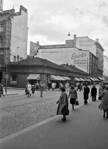 Alexandersgatan 9. - Glogatan 5. Ett lågt trähus omringat av högre stenhus. Fotgängare på båda sidor av gatan.