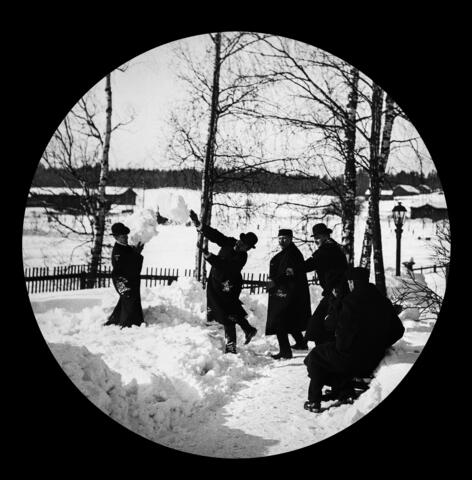 Män klädda i hattar och långa kappor leker med snöbollar. Bakom dem finns björkar och en gaslykta.