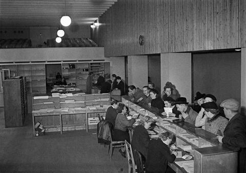 Työntekijöiden puolelta tiskiä otetussa valokuvassa näkyy työntekijöitä takaa sivuttain ja asiakkaita tiskin toisella puolella edestä sivuttain. 