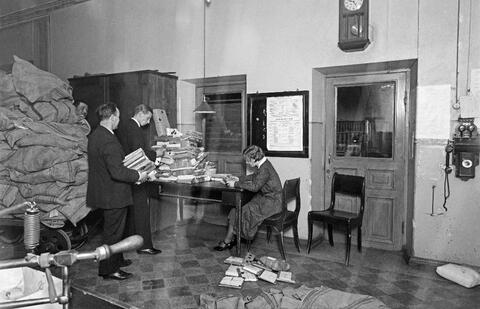 Till vänster en transportkärra med en stor hög säckar ovanpå. Två män bär paketen på ett bord som redan har stora pappershögar. Vid bordet sitter en kvinna med ett paket i handen. Det finns paket också på golvet. 