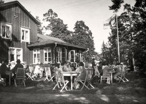 Människor sitter utanför en kafébyggnad på sommaren vid små runda bord. Finska flaggan vajar i flaggstången.