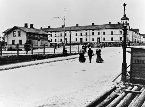 Ljusa tvåvåningsbyggnader, en man omgiven av tre kvinnor promenerar bortåt från tittaren, till höger en gaslykta.