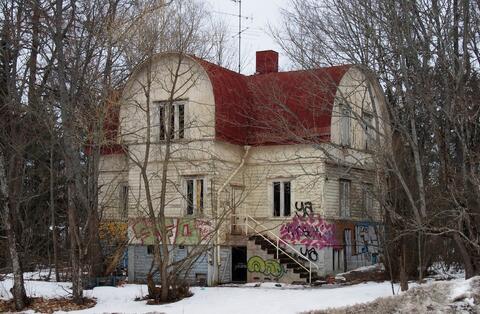 Kaksikerroksinen valkoinen puuhuvila, jossa on punainen katto. Talon ulkoseiniin on maalattu graffiteja ja julkisivu on huonossa kunnossa. 