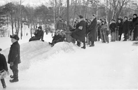 Ett snöigt landskap med vuxna och barn som samlats på ett backkrön med pulkor och kälkar. Två barn är på väg att åka, de andra väntar på sin tur. 
