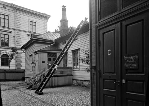 Oikealla Helsingin ruotsalais-suomalaisen seurakunnan pastorinkanslian sisäänkäynti. Vasemmalla sisäänkäynti Kristliga Föreningen av Unga Män (Nuorten miesten kristillinen yhdistys) -yhdistyksen tiloihin. Katolle vieviin tikkaisiin nojaa mahdollisen tulipalon sammuttamisessa käytettävä palohaka. Taustalla on Svenska Fruntimmerskolanin eli ruotsalaisen tyttökoulun rakennus osoitteessa Bulevardi 18–20, jossa nykyään toimii Helsingin eurooppalainen koulu. Vuodelta 1885 olevan koulurakennuksen suunnittelijat ov