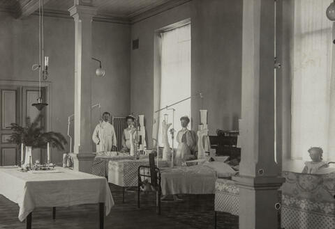  Kuvassa on kaksi sairaanhoitajaa ja lääkäri sekä neljä lapsipotilasta, joilla kaikilla on toinen alaraaja vetolaitteessa. Lisäksi kuvan oikeassa reunassa on sairaalassa vierailulla oleva tyttö.