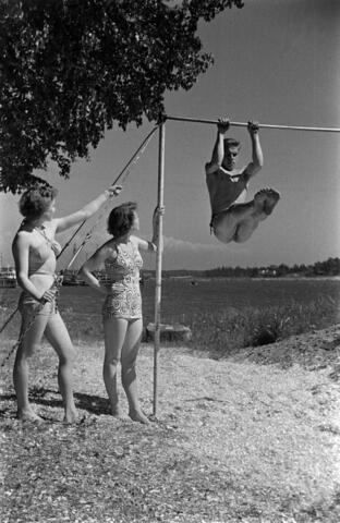 Kaksi naista ja mies Sompasaaren rannalla. Mies voimistelee rekkitangolla.