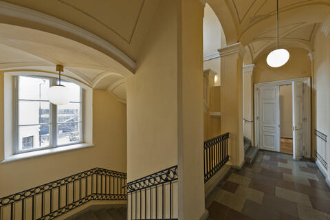 A light-coloured stairwell with a grey stone floor, an arched ceiling and spherical ceiling lights. On the right is a corridor with white double doors at the end, one of which is open.