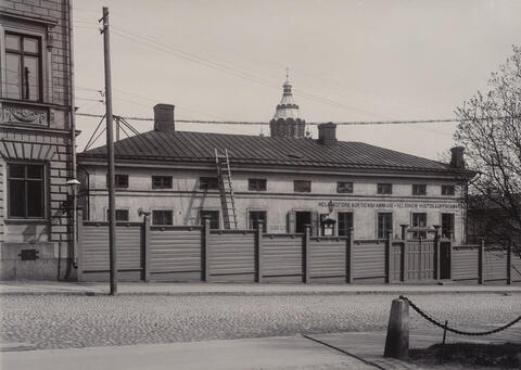 Behind the board fence is a mansard-roofed two-storey building, with the tower of Uspenski Cathedral looming behind.