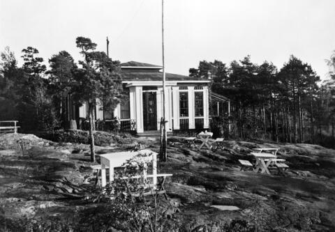 Ett envånings trähus omgiven av unga tallar med en flaggstång framför.
