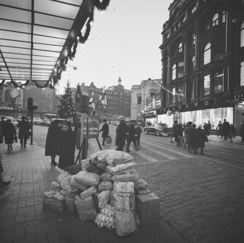 Pelastusarmeijan joulupata Stockmannin tavaratalon edessä Helsingissä. Etualalla on kasa paketteja ja niiden takana pata, taustalla on jalankulkijoita.