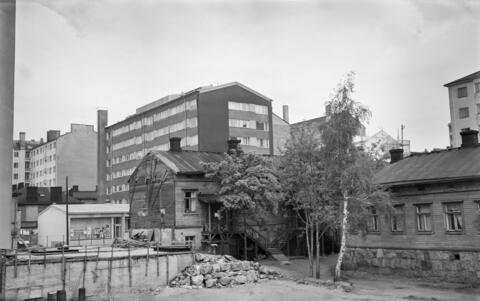 I förgrunden ser man en tom gård och lövträd, och bakom dem låga trähus. Nya flervåningshus reser sig i bakgrunden. 