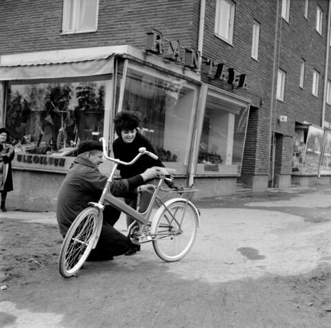 Etualalla on polkupyörä, jonka takana hymyilee nuori nainen. Vasemmalla on mies kyykistyneenä säätämään pyörän satulaa. Taustalla on punatiilirakennus, jonka näyteikkunoissa on polkupyöriä. Näyteikkunan yläpuolella on valokirjaimet RANTALA.