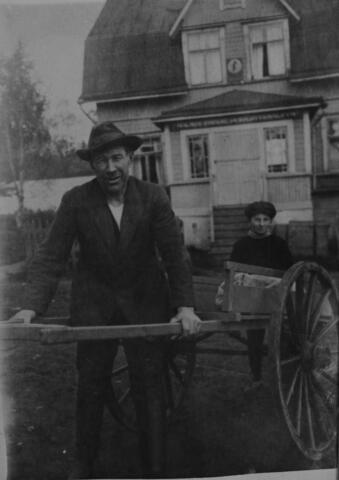 En småleende man i hatt och vardagskostym håller i skaklarna av en enkel kärra av trä. Bakom honom syns en stor säck på kärran och en pojke i keps. I bakgrunden står ett trähus med valmtak 