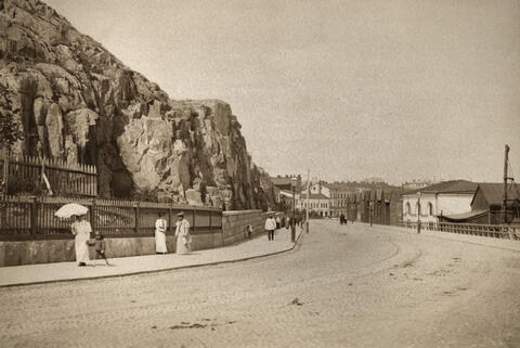 Ihmisiä kävelee rantakadulla. Vasemmalla on kallionseinämää, oikealla on satamamakasiineja.