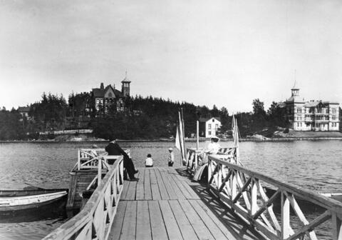 En träbrygga vid Tölövikens strand. En man och kvinna sitter på bänkarna vid sidorna av bryggan. I ändan av bryggan står tre barn. På andra stranden två trävillor. 