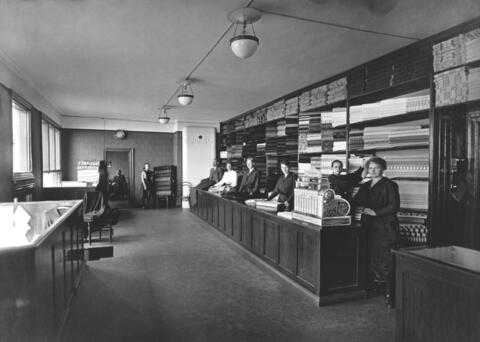 Pitkänomainen myymälätila, jossa on myyntitiskin takana kuusi myyjää ja kangaspakkoja seinähyllyissä. Sivuseinällä on kangaspinon vieressä kauppa-apulainen ja nurkassa kaakeliuuni. Myymälähuoneen takana näkyy kauppias Ymär Abdrahim toimistossaan.