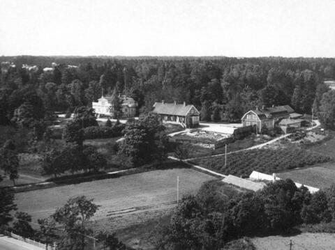 Etualalla näkyy peltoja, kasvihuoneita ja puutarhaa, keskellä näkyy kolme maatilan rakennusta ja taustalla metsää.