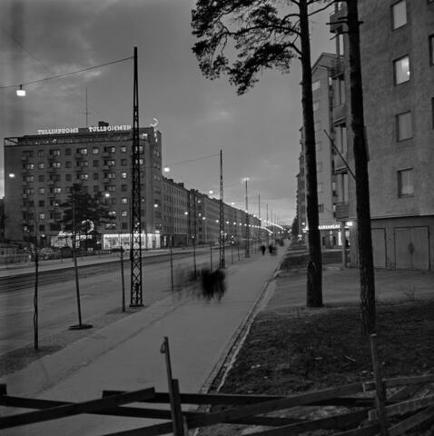 Näkymä Mannerheimintietä luoteeseen. Edessä oikealla Mannerheimintie 73 kulma, jonka jälkeen Mannerheimintie 75. Vasemmalla Mannerheimintie 118, ns. Tullinpuomin talo, jossa ravintola Tullinpuomi ja elokuvateatteri Sininen kuu. Jalkakäytävä on sateen kastelema, ja siinä on jokunen jalankulkija. 