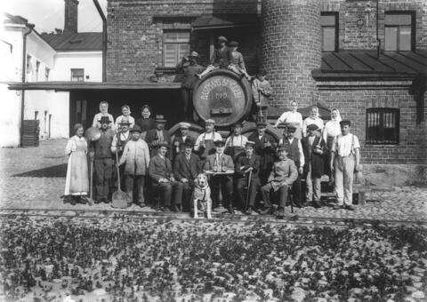 Noin 30 ihmistä kokoontuneena punatiilirakennuksen eteen. Taustalla on oluttynnyreitä, joiden päällä istuu kaksi poikaa ja seisoo kaksi miestä.