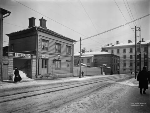 Vasemmalla on kaksikerroksiset puutalot ja puinen piha-aita. Avoimesta pihaportista näkyy lähes ihmisen korkuinen lumikasa. Jalkakäytävällä kävelee viisi ihmistä. Taustalla on Kaartin kasarmi.