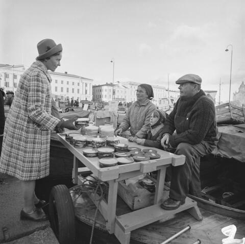 En man, en kvinna och ett barn sitter tanför en båt bakom ett bord fyllt med strömmingsburkar. På andra sidan bordet står en kvinna i rutig kappa. Hon håller en fiskburk i handen.