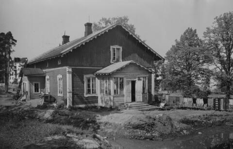 Ett trähus på en och en halv våning med öppna pardörrar. På gården finns en lina med byk på tork.