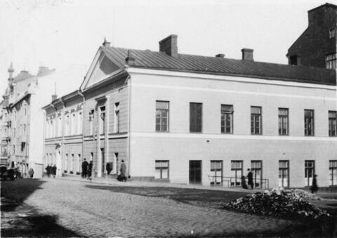 Kadunkulmassa näkyy kaksikerroksinen kivitalo, jonka katolla on savupiippuja. 