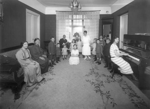 Aikuisia ja lapsia oleskeluhuoneessa. Nuori nainen istuu pianon ääressä.