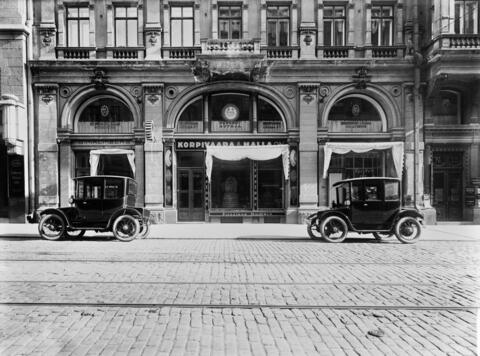 Etualalla on mukulakivikatua, jossa raitiovaunukiskot, taka-alalla rakennuksen julkisivu. Kadulle rakennuksen eteen on pysäköity kaksi autoa.