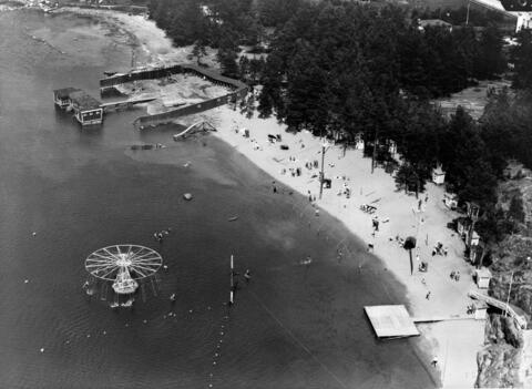 Flygbild från en sandstrand med vattenkarusell, badande människor och omklädningshytter.