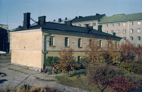 En gul tvåvånings byggnad med barn vid hörnet och lövträd i höstdräkt framför. Bakom huset syns gråa nyare höghus.