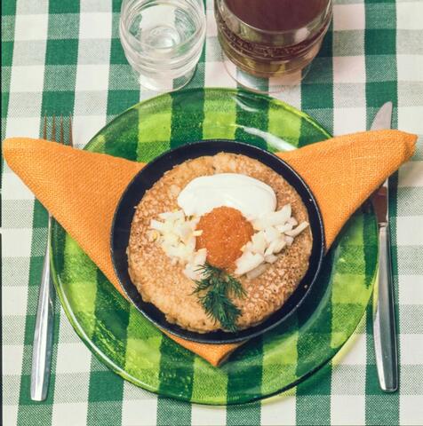 Närbild från ovan av en blini med lök, smetana, fiskrom och dill. Blinin är placerad i ett kärl av metall ovanpå en grön glastallrik och en orange tygservett. Bordet är dukat med en grönvitrutig tygduk, bestick samt ett glas vatten och ett glas svagdricka.