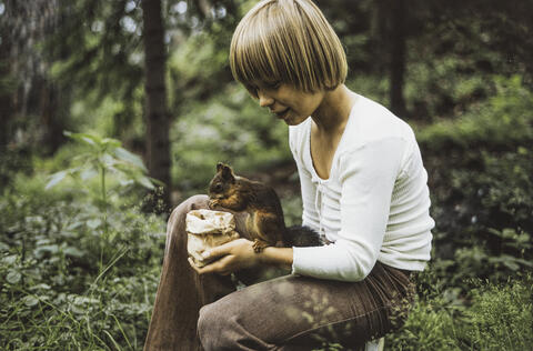 Hymyilevä poika on polvistunut maahan. Hänellä on valkoinen paita ja ruskeat vakosamettihousut. Hänen käsivarrellaan istuu orava, joka syö hänen kädessään olevasta paperipussista.