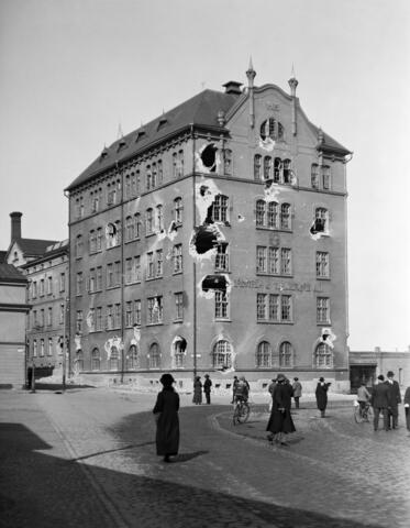 Kuvan keskellä on vaurioitunut viisikerroksinen tehdasrakennus, etualalla ihmisiä kävelemässä ja pyöräilemässä. 