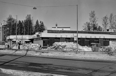 Månsas köpcentrum, Baggbölevägen 11. Till vänster i bakgrunden syns Baggbölevägen 13. Fotografiet har tagits på vårvintern. Gatan kantas av stora snödrivor. Till höger syns en korvkiosk framför köpcentret.