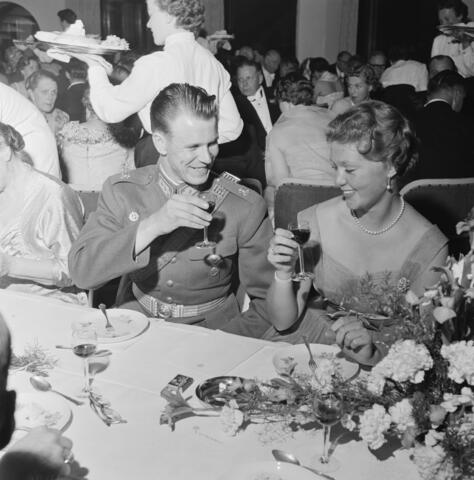 : Svenska prinsessorna Sibylla, Desiré och Birgitta på besök i Finland. Festmiddag som arrangerats av Finland automobilklubb på Fiskartorpet. Prinsessan Birgitta sitter vid middagsbordet och skålar med en officer som sitter bredvid. I bakgrunden syns en servitör och andra middagsgäster.