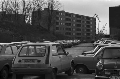 I förgrunden står en massa bilar parkerade. Bakom dem tornar sig ett sexvåningars elementhus. En lyftkran skymtar bakom huset.