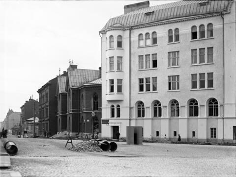 Katunäkymä Fredrikinkadun ja Malminkadun risteyksestä. Autiolla tiellä on viemäriputkia odottamassa asennusta. Ensimmäinen punatiilirakennus oikealta on Luther-kirkko.