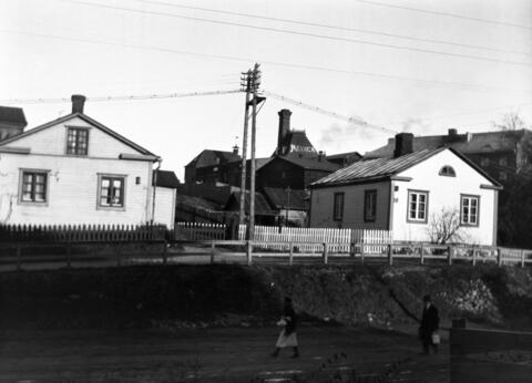 Låga ljusa trähus vid en ladsväg. I mitten i bakgrunden en fabriksbyggnad i rödtegel.