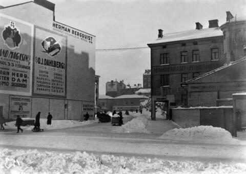 Talojen välissä kulkee uusi katu, jolla ajaa autoja ja jonka sivuilla on ihmisiä. Maassa on paljon lunta. 