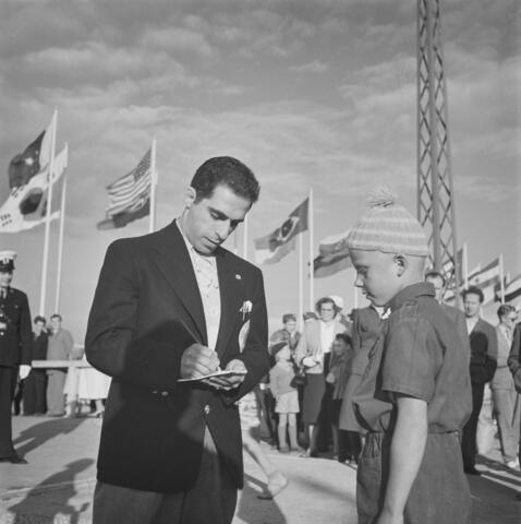 A male athlete is writing his autograph, while a young boy waits