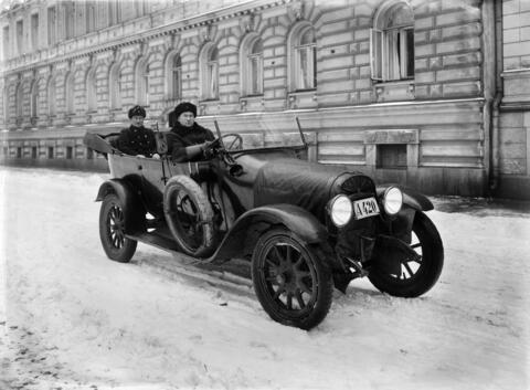 Avoauto, jonka takapenkillä kuljettajan takana istuu mies, jolla on univormumainen takki ja karvalakki. Kuljettajalla on talvitakin ja -hatun lisäksi nahkaiset ajurinhansikkaat. Auton ohjauspyörä on vasemmalla. Maassa on lunta, ja taustalla näkyy leveä empire-julkisivu.