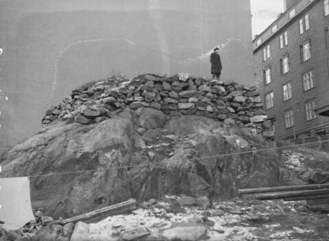I bakgrunden en husvägg, intill den på innergården ett berg med stenröse ovanpå. En människa står uppe på det.