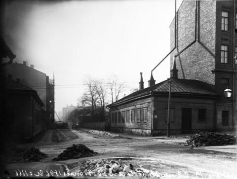 Keskellä kuvaa on matala puutalo, jonka oikealla puolella kohoaa korkea tiilitalo. Vasemmalla näkyy matala puutalo ja kaksikerroksinen kivitalo.