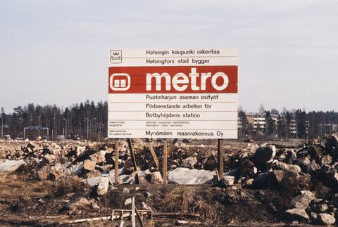 Kivilouhikon edustalla rakennustyömaan infokyltti: "Helsingin kaupunki rakentaa - Metro - Puotinharjun aseman esityöt - Mynämäen maanrakennus oy". Kyltissä myös metron vanhempi, siankärsäksi kutsuttu tunnelilogo.